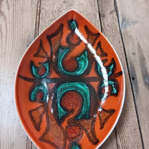 Könnte beinhalten: Eine orangefarbene, blattförmige Keramikschale mit weißem Rand. Die Schale hat ein auffälliges, abstraktes Design in Grün-, Braun- und Rottönen. Das Design ist symmetrisch und scheint handgemalt zu sein. Die Schale ist wahrscheinlich Vintage oder Retro.