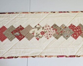 May include: A white table runner with a diamond pattern made of red, brown, and cream fabric. The runner has a floral print and a pink and brown border.
