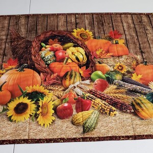 May include: A rectangular placemat featuring a cornucopia overflowing with pumpkins, gourds, apples, and corn. Sunflowers and fall leaves surround the harvest scene, set against a wooden background. The border has a brown floral pattern.