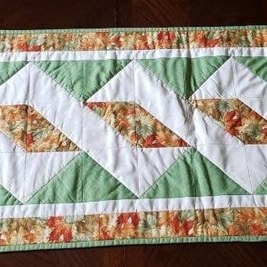 May include: A rectangular quilted table runner with a repeating geometric pattern. The design features white, green, and autumn-colored floral fabric. The runner is approximately 12 inches wide and 40 inches long.
