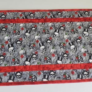 May include: Rectangular quilted table runner featuring a repeating pattern of skulls with top hats and roses on a gray background. The runner has a red border and is likely handmade.