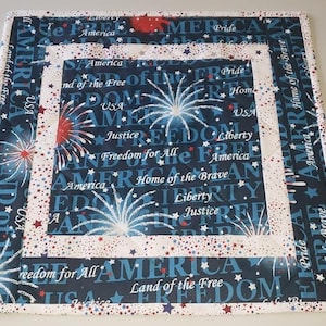 May include: A blue and white fabric placemat with a red, white, and blue patriotic design. The placemat features fireworks, stars, and the words "America", "Land of the Free", "Home of the Brave", "Freedom for All", "Liberty", and "Justice".