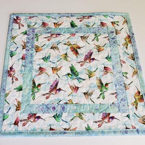 May include: Square quilted placemat featuring a hummingbird pattern in shades of green, purple, orange, and blue on a white background. The placemat has a light blue border and a quilted design, perfect for a dining table.