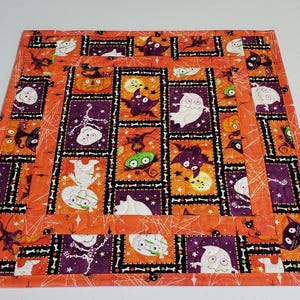 May include: Square Halloween-themed fabric placemat. The orange background features a repeating pattern of ghosts, bats, pumpkins, and skeletons in purple and orange squares. The placemat has a stitched border.