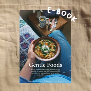 Puede incluir: Portada de un libro electrónico titulado "Gentle Foods" con un cuenco de sopa con crema. El texto describe recetas veganas, sin gluten y bajas en FODMAP. El fondo incluye una mesa con una taza y una manta estampada.