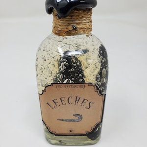 Può includere: Una bottiglia da farmacista in vetro trasparente con un sigillo di cera nero e un'etichetta che recita "Leeches". La bottiglia contiene oggetti scuri a forma di verme ed è avvolta con spago attorno al collo. L'etichetta dice anche "CSD Apothecary".