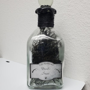 May include: A clear glass bottle with a black stopper and a label that reads "CSD Apothecary Devil's Snare". The bottle is filled with dried black herbs.