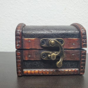 Op de afbeelding: Een kleine houten schatkist met een donkerbruine, getextureerde afwerking en lichtere bruine randen. De kist heeft een metalen sluiting en scharnieren. Geschikt voor het bewaren van kleine spullen of sieraden.