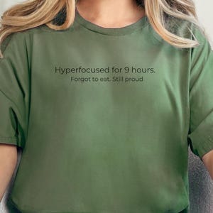 May include: An olive green t-shirt with the text "Hyperfocused for 9 hours. Forgot to eat. Still proud." The shirt has short sleeves and a relaxed fit. The model is wearing light blue ripped jeans.