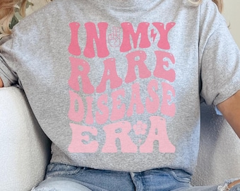 May include: A gray t-shirt with a pink graphic that says "In My Rare Disease Era" in a retro font.
