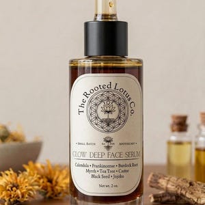 Op de afbeelding: Een heldere glazen fles "Glow Deep Face Serum" met een zwarte dop en pipet. Het etiket toont "The Rooted Lotus Co." en een lijst met ingredi&euml;nten. De fles bevat een bruine vloeistof. Nettogewicht 57 g.