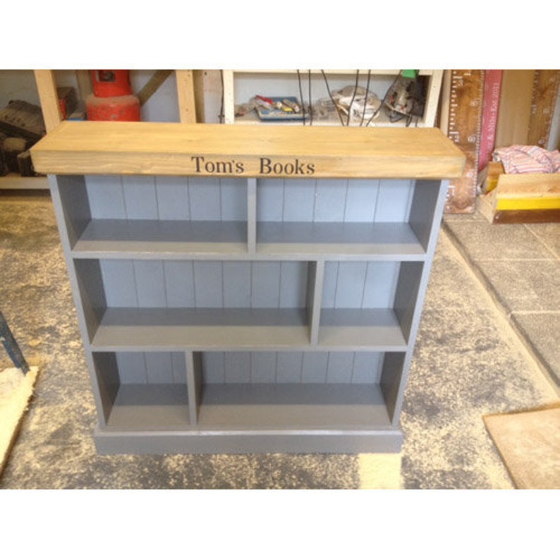 May include: A gray wooden bookshelf with a light wood top. The top has the words "Tom's Books" engraved on it. The bookshelf has multiple shelves and compartments.