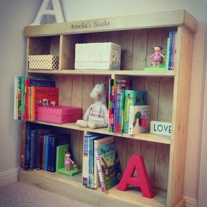 May include: A wooden bookshelf with multiple shelves, filled with books and toys. The top shelf has the words "Amelia's Books" written on it. The bookshelf is decorated with a large letter A and a small doll.