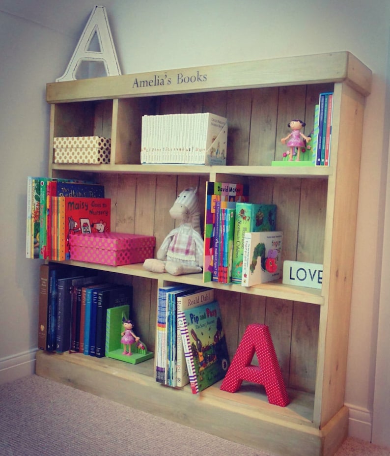 May include: A wooden bookshelf with multiple shelves, filled with books and toys. The top shelf has the words "Amelia's Books" written on it. The bookshelf is decorated with a large letter A and a small doll.