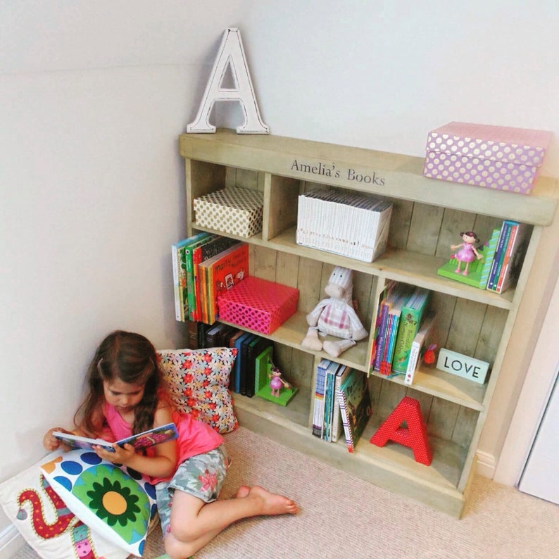 May include: A wooden bookshelf with five shelves, filled with books and decorative items. The bookshelf is in a corner of a room and has the words "Amelia's Books" written on the top shelf. A young girl is sitting on the floor in front of the bookshelf, reading a book.
