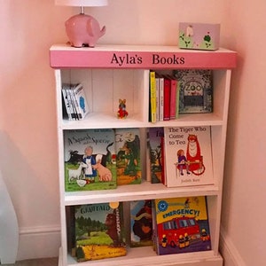 May include: A white bookshelf with pink trim and the words "Ayla's Books" on the top shelf. The bookshelf is filled with children's books, including "The Tiger Who Came to Tea", "Gruffalo", and "Emergency!"
