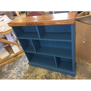 May include: A blue wooden bookcase with a wood top and the words "Alfie's Books" engraved on the front. The bookcase has multiple shelves and compartments.