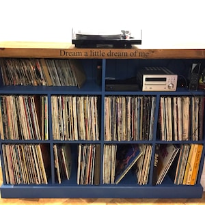 May include: A blue record player cabinet with a wooden top. The cabinet has 12 shelves for storing vinyl records. The top of the cabinet has a turntable and a stereo receiver. The cabinet has the text "Dream a little dream of me" engraved on the top.