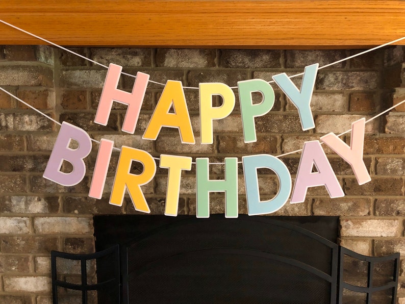 HAPPY BIRTHDAY banner made with pastel cardstock. Background made from white cardstock. Letters are cut out of different colors of sturdy cardstock. These are attached with 3d tape. Letters are strung together with white ribbon. There is extra ribbon on the ends for you to hang it up.