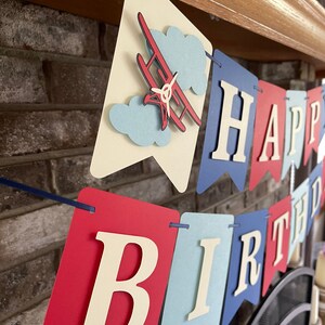 Vintage airplane Happy Birthday banner. Alternating navy blue, red and light blue cardstock flags. Ivory letters are attached with 3D tape. 3D airplanes decorate the two end flags. The banner is strung on a matching ribbon ready to hang.
