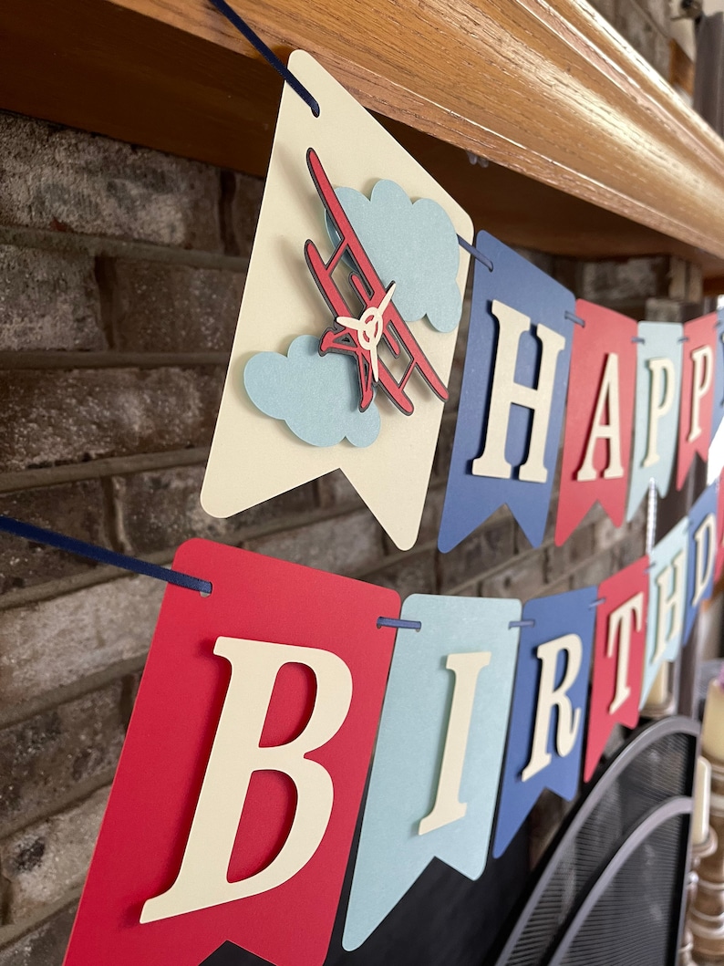 Vintage airplane Happy Birthday banner. Alternating navy blue, red and light blue cardstock flags. Ivory letters are attached with 3D tape. 3D airplanes decorate the two end flags. The banner is strung on a matching ribbon ready to hang.