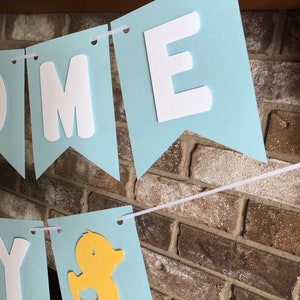 Rubber duck Welcome Baby sign. The flags are made with light blue cardstock backgrounds and white letters. The letters are attached with 3d tape to give it dimension. The two end flags are decorated with yellow rubber ducks. These are strung on a coordinating satin ribbon with plenty of extra to hang the banner.