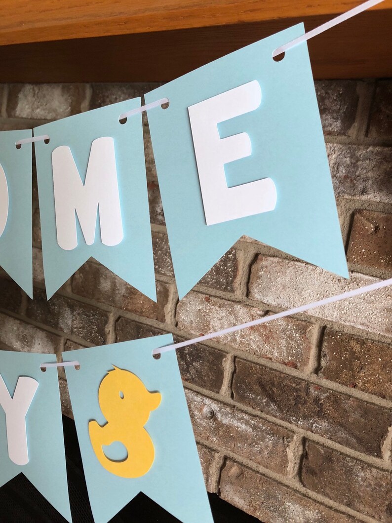 Rubber duck Welcome Baby sign. The flags are made with light blue cardstock backgrounds and white letters. The letters are attached with 3d tape to give it dimension. The two end flags are decorated with yellow rubber ducks. These are strung on a coordinating satin ribbon with plenty of extra to hang the banner.