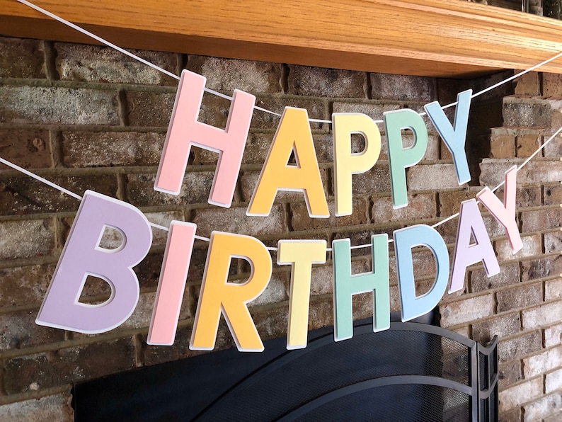HAPPY BIRTHDAY banner made with pastel cardstock. Background made from white cardstock. Letters are cut out of pastel colors of sturdy cardstock. These are attached with 3d tape. Letters are strung together with white ribbon. There is extra ribbon on the ends for you to hang it up.