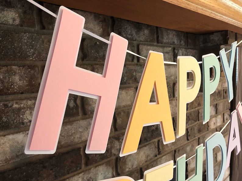 HAPPY BIRTHDAY banner made with pastel cardstock. Background made from white cardstock. Letters are cut out of different colors of sturdy cardstock. These are attached with 3d tape. Letters are strung together with white ribbon. There is extra ribbon on the ends for you to hang it up.
