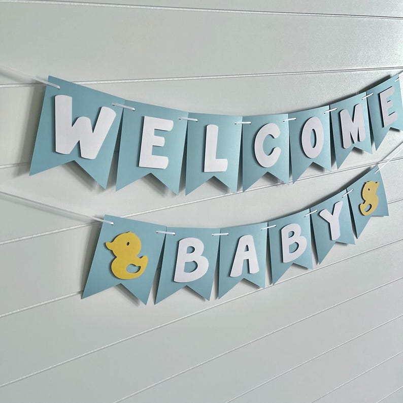 Rubber duck Welcome Baby sign. The flags are made with light blue cardstock backgrounds and white letters. The letters are attached with 3d tape to give it dimension. The two end flags are decorated with yellow rubber ducks. These are strung on a coordinating satin ribbon with plenty of extra to hang the banner.
