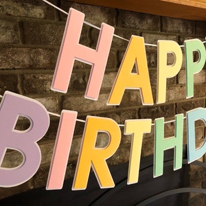 HAPPY BIRTHDAY banner made with pastel cardstock. Background made from white cardstock. Letters are cut out of different colors of sturdy cardstock. These are attached with 3d tape. Letters are strung together with white ribbon. There is extra ribbon on the ends for you to hang it up.