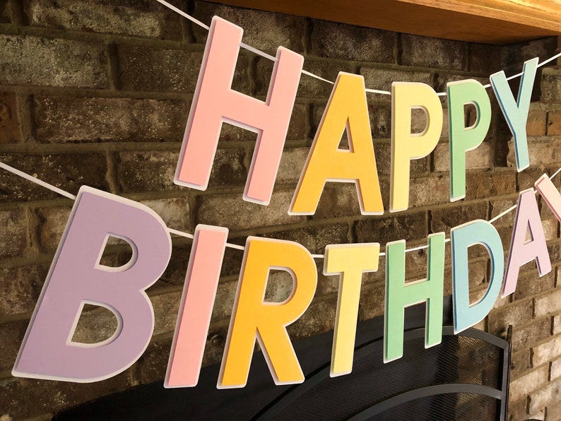 HAPPY BIRTHDAY banner made with pastel cardstock. Background made from white cardstock. Letters are cut out of different colors of sturdy cardstock. These are attached with 3d tape. Letters are strung together with white ribbon. There is extra ribbon on the ends for you to hang it up.