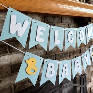 Rubber duck Welcome Baby sign. The flags are made with light blue cardstock backgrounds and white letters. The letters are attached with 3d tape to give it dimension. The two end flags are decorated with yellow rubber ducks. These are strung on a coordinating satin ribbon with plenty of extra to hang the banner.