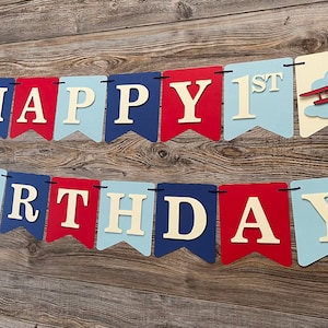 Vintage airplane Happy Birthday banner. Alternating navy blue, red and light blue cardstock flags. Ivory letters are attached with 3D tape. 3D airplanes decorate the two end flags. The banner is strung on a matching ribbon ready to hang.