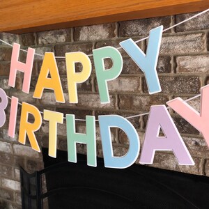 HAPPY BIRTHDAY banner made with pastel cardstock. Background made from white cardstock. Letters are cut out of different colors of sturdy cardstock. These are attached with 3d tape. Letters are strung together with white ribbon. There is extra ribbon on the ends for you to hang it up.