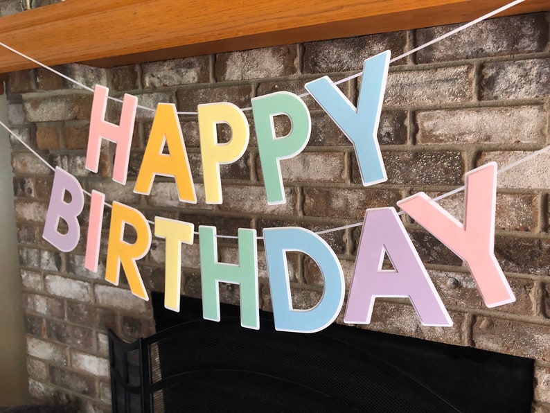 HAPPY BIRTHDAY banner made with pastel cardstock. Background made from white cardstock. Letters are cut out of different colors of sturdy cardstock. These are attached with 3d tape. Letters are strung together with white ribbon. There is extra ribbon on the ends for you to hang it up.