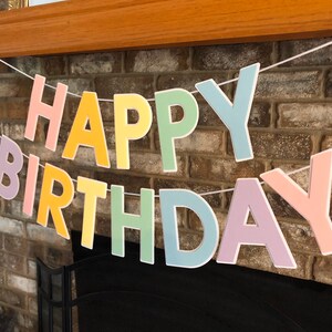 HAPPY BIRTHDAY banner made with pastel cardstock. Background made from white cardstock. Letters are cut out of different colors of sturdy cardstock. These are attached with 3d tape. Letters are strung together with white ribbon. There is extra ribbon on the ends for you to hang it up.