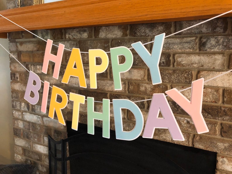 HAPPY BIRTHDAY banner made with pastel cardstock. Background made from white cardstock. Letters are cut out of different colors of sturdy cardstock. These are attached with 3d tape. Letters are strung together with white ribbon. There is extra ribbon on the ends for you to hang it up.
