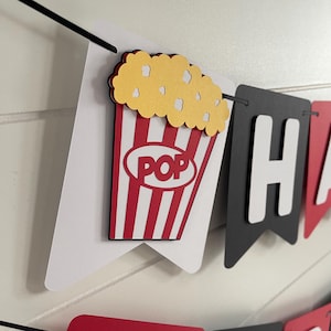 May include: A banner with a popcorn bucket design. The popcorn bucket is red and white striped with the word "POP" in a white oval. The popcorn is yellow. The banner also has black and red pennants.