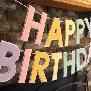 HAPPY BIRTHDAY banner made with pastel cardstock. Background made from white cardstock. Letters are cut out of different colors of sturdy cardstock. These are attached with 3d tape. Letters are strung together with white ribbon. There is extra ribbon on the ends for you to hang it up.