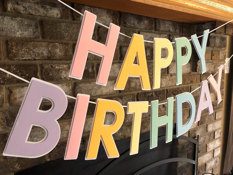 HAPPY BIRTHDAY banner made with pastel cardstock. Background made from white cardstock. Letters are cut out of different colors of sturdy cardstock. These are attached with 3d tape. Letters are strung together with white ribbon. There is extra ribbon on the ends for you to hang it up.