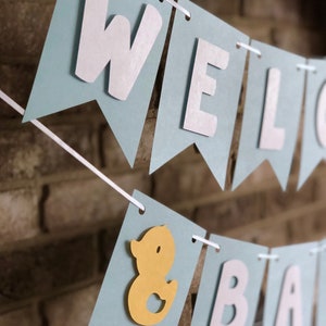 Rubber duck Welcome Baby sign. The flags are made with light blue cardstock backgrounds and white letters. The letters are attached with 3d tape to give it dimension. The two end flags are decorated with yellow rubber ducks. These are strung on a coordinating satin ribbon with plenty of extra to hang the banner.