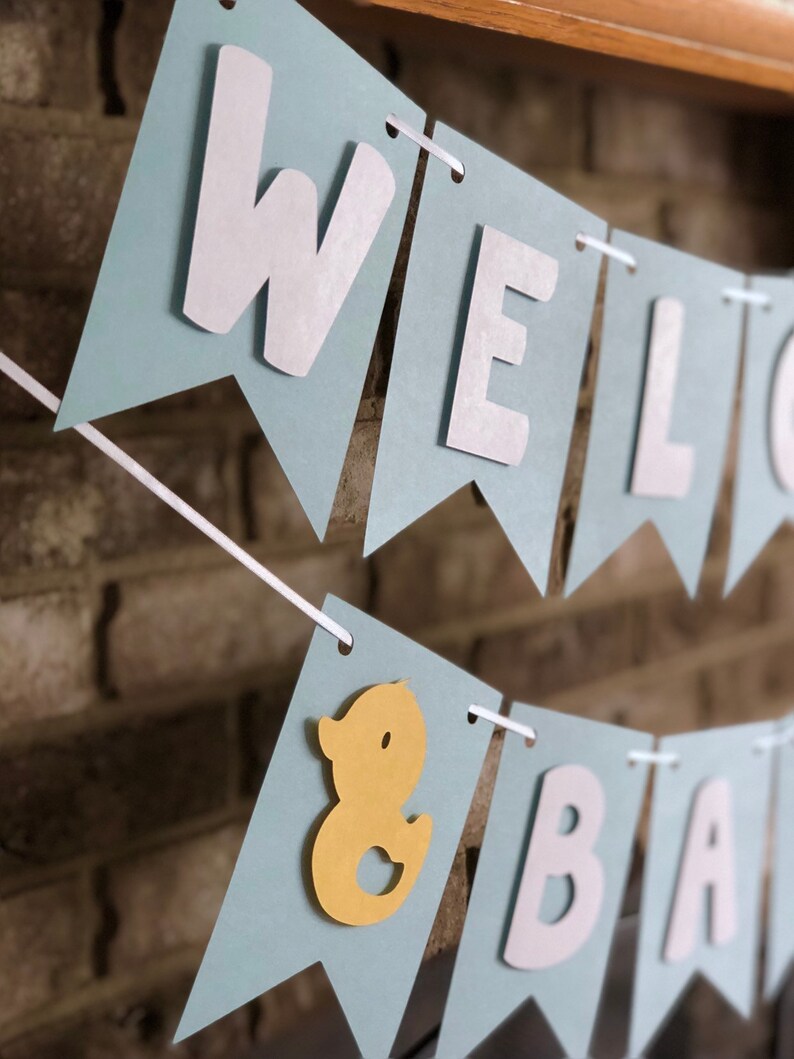 Rubber duck Welcome Baby sign. The flags are made with light blue cardstock backgrounds and white letters. The letters are attached with 3d tape to give it dimension. The two end flags are decorated with yellow rubber ducks. These are strung on a coordinating satin ribbon with plenty of extra to hang the banner.