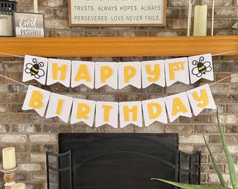 May include: A white banner with yellow letters spelling "HAPPY 1ST BIRTHDAY" is strung across a fireplace. The banner has bee graphics on each end. The background includes a brick fireplace, candles, and decorative items.