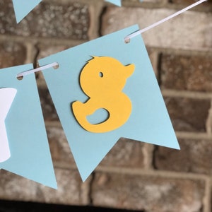 Rubber duck Welcome Baby sign. The flags are made with light blue cardstock backgrounds and white letters. The letters are attached with 3d tape to give it dimension. The two end flags are decorated with yellow rubber ducks. These are strung on a coordinating satin ribbon with plenty of extra to hang the banner.