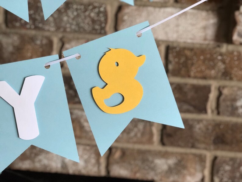 Rubber duck Welcome Baby sign. The flags are made with light blue cardstock backgrounds and white letters. The letters are attached with 3d tape to give it dimension. The two end flags are decorated with yellow rubber ducks. These are strung on a coordinating satin ribbon with plenty of extra to hang the banner.