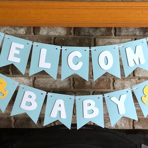 Rubber duck Welcome Baby sign. The flags are made with light blue cardstock backgrounds and white letters. The letters are attached with 3d tape to give it dimension. The two end flags are decorated with yellow rubber ducks. These are strung on a coordinating satin ribbon with plenty of extra to hang the banner.