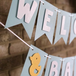 Rubber duck Welcome Baby sign. The flags are made with light blue cardstock backgrounds and white letters. The letters are attached with 3d tape to give it dimension. The two end flags are decorated with yellow rubber ducks. These are strung on a coordinating satin ribbon with plenty of extra to hang the banner.