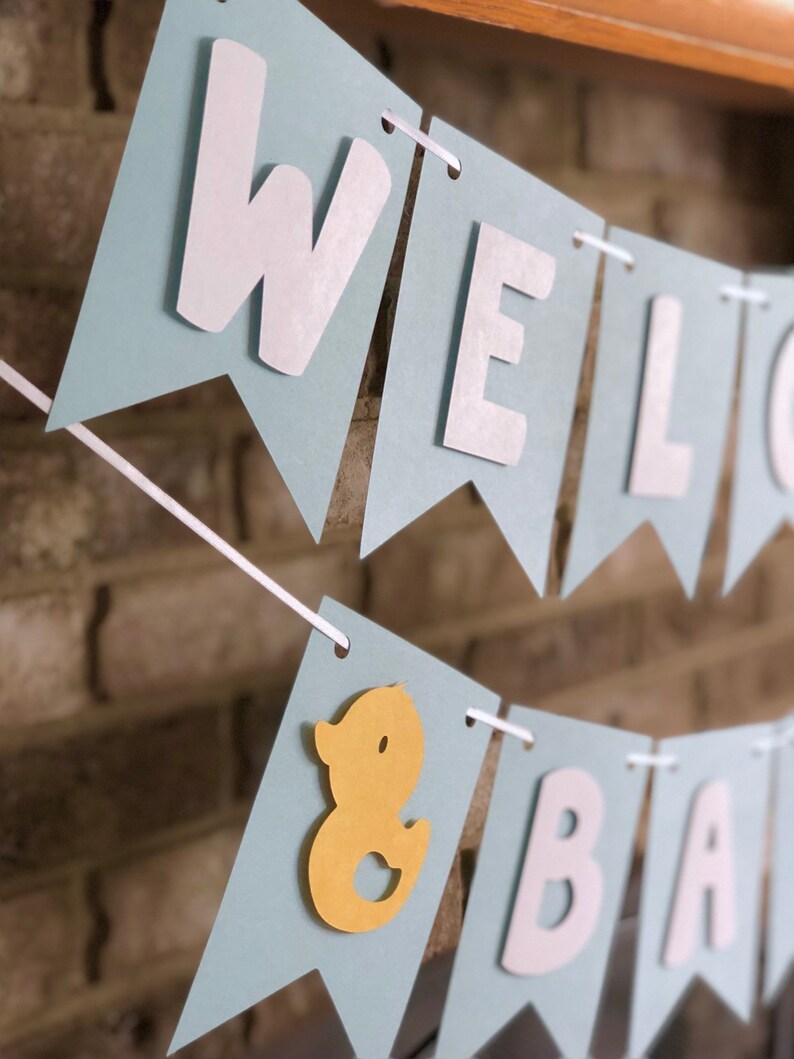 Rubber duck Welcome Baby sign. The flags are made with light blue cardstock backgrounds and white letters. The letters are attached with 3d tape to give it dimension. The two end flags are decorated with yellow rubber ducks. These are strung on a coordinating satin ribbon with plenty of extra to hang the banner.