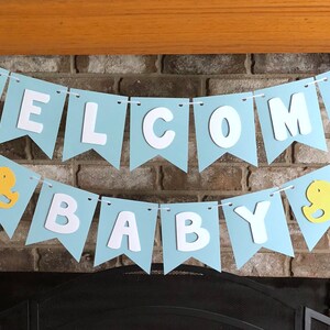 Rubber duck Welcome Baby sign. The flags are made with light blue cardstock backgrounds and white letters. The letters are attached with 3d tape to give it dimension. The two end flags are decorated with yellow rubber ducks. These are strung on a coordinating satin ribbon with plenty of extra to hang the banner.